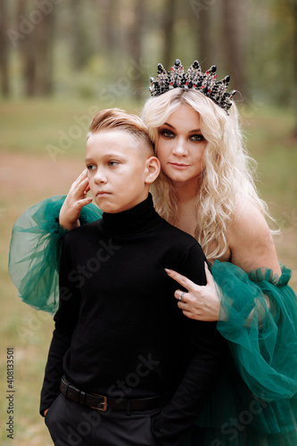 young beautiful blonde woman queen with young boy in black outfit. Princess mother walks with son. autumn green forest mystic. Vintage medieval shiny crown. Long evening green dress. magic fantasy