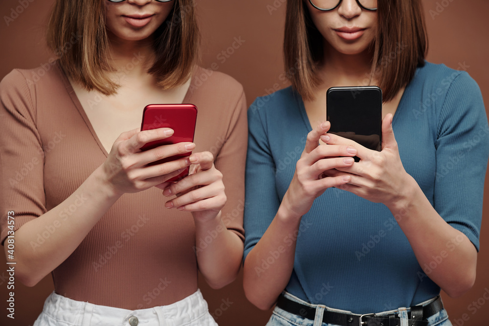 Brunette twin sisters standing close to one another and scrolling in smartphones Brunette twin sisters standing close to one another and scrolling in smartphones