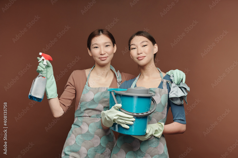Happy young female twins in aprons and gloves holding detergent and bucket