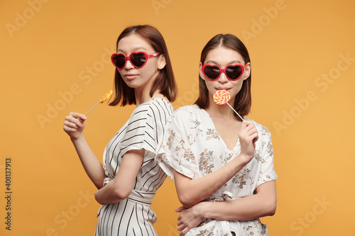Young stylish female twins in smart dresses and sunglasses having lollipops