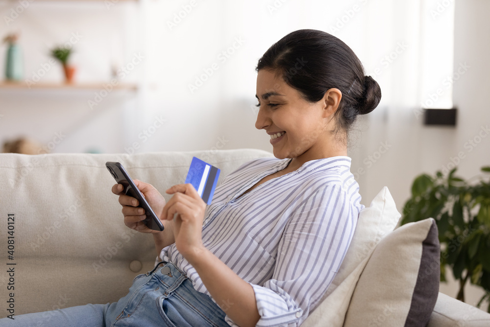 Young indian woman shopper sit on couch enjoy easy paying for goods online using bank card phone. Happy mixed race female traveler book tickets trip hotel at travel agency transport operator website Young indian woman shopper sit on couch enjoy easy paying for goods online using bank card phone. Happy mixed race female traveler book tickets trip hotel at travel agency transport operator website