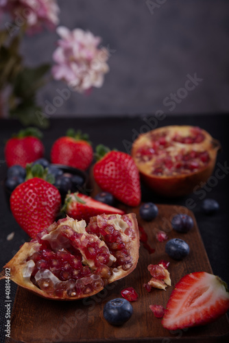 Strawberries, blueberries and pomegranates exposed on a wooden board in dark background.
