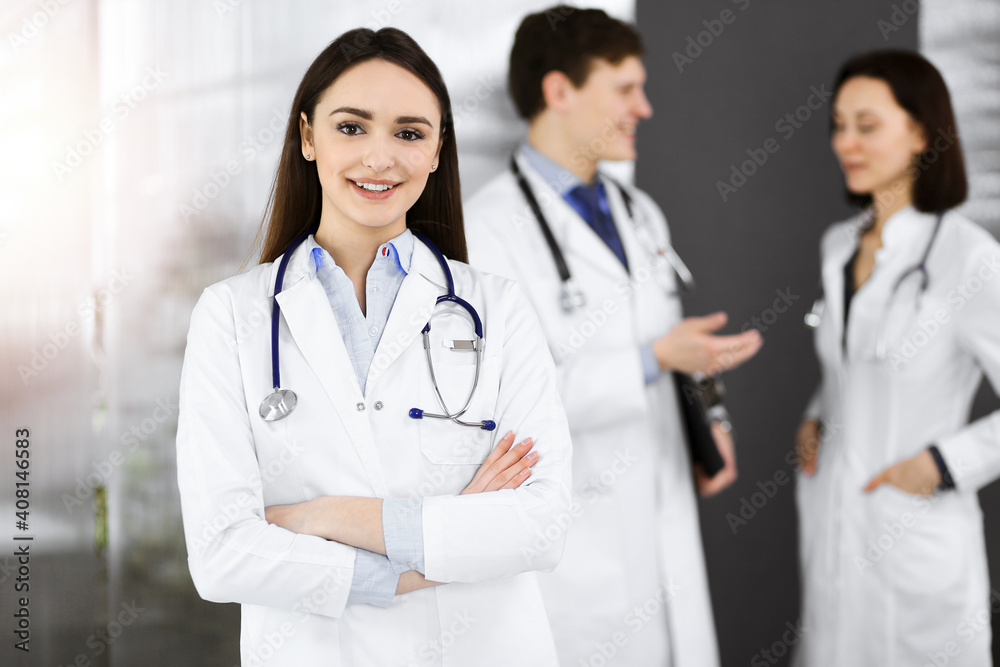 Smiling intelligent woman-doctor is standing with arms crossed in sunny clinic together with her colleagues at the background. Perfect medical service in a hospital Smiling intelligent woman-doctor is standing with arms crossed in sunny clinic together with her colleagues at the background. Perfect medical service in a hospital