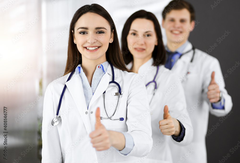 Group of young cheerful doctors standing as a team with thumbs up in sunny hospital. Medical help, insurance in health care, best disease treatment and medicine concept Group of young cheerful doctors standing as a team with thumbs up in sunny hospital. Medical help, insurance in health care, best disease treatment and medicine concept