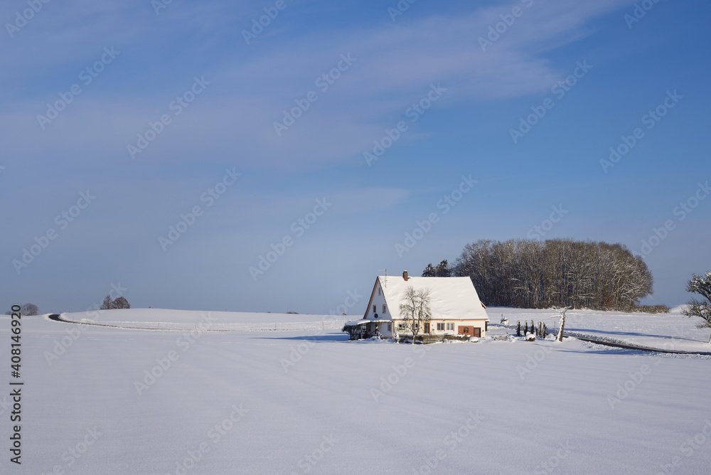 Winter landscape in Bavaria with a house that is covered in snow, behind a blanket of snow, in front of bare trees and a blue sky in winter Winter landscape in Bavaria with a house that is covered in snow, behind a blanket of snow, in front of bare trees and a blue sky in winter