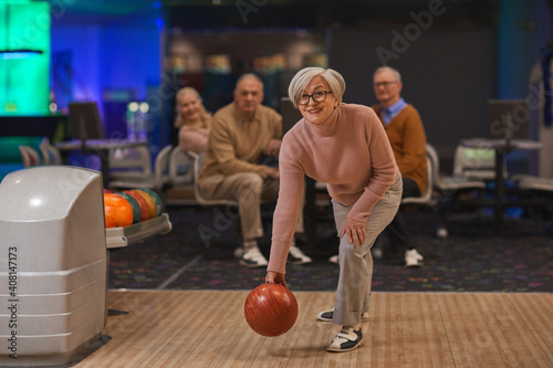 Full length portrait of joyful senior woman playing bowling with group of friends in background, copy space