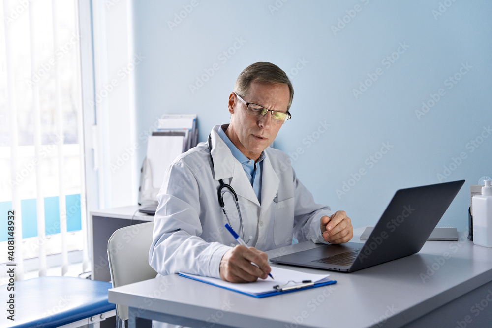 Serious old mature professional male doctor using laptop computer in hospital office watching medical webinar training, writing in healthcare report, consulting patient online at telemedicine meeting.