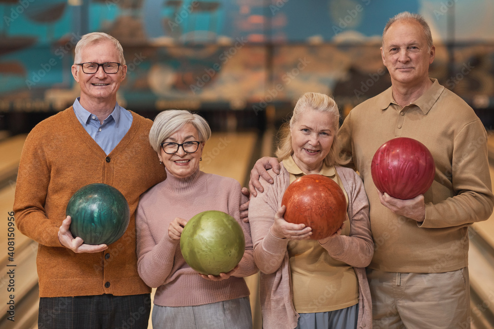 Fototapeta premium Waist up portrait of group of happy senior people holding bowling balls and smiling at camera while enjoying active entertainment at bowling alley, copy space