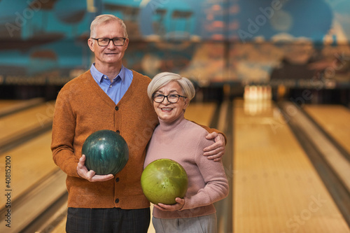 Waist up portrait of happy senior couple holding bowling balls and smiling at camera while enjoying active entertainment at bowling alley, copy space