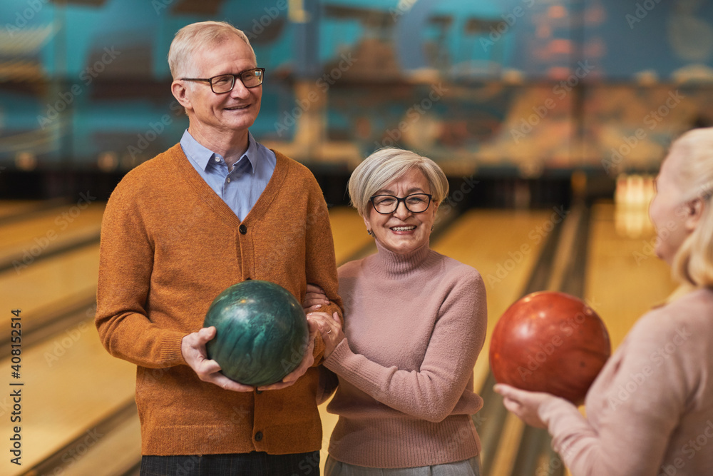 Fototapeta premium Waist up portrait of group of happy senior people holding bowling balls and chatting while enjoying active entertainment at bowling alley, copy space