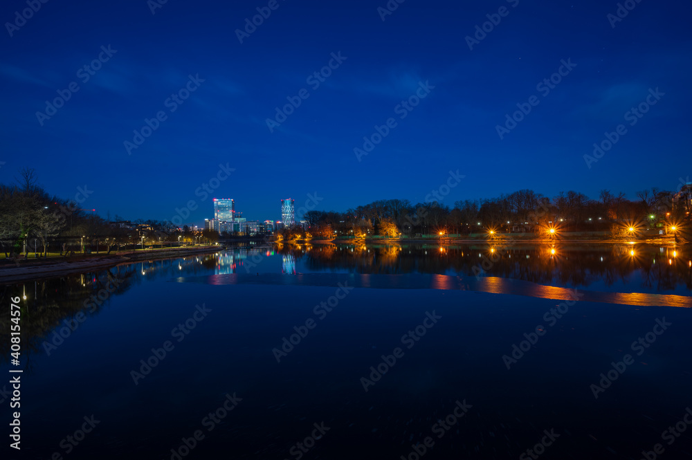 Lake in park during blue hour with illuminated office buildings and apartment blocks