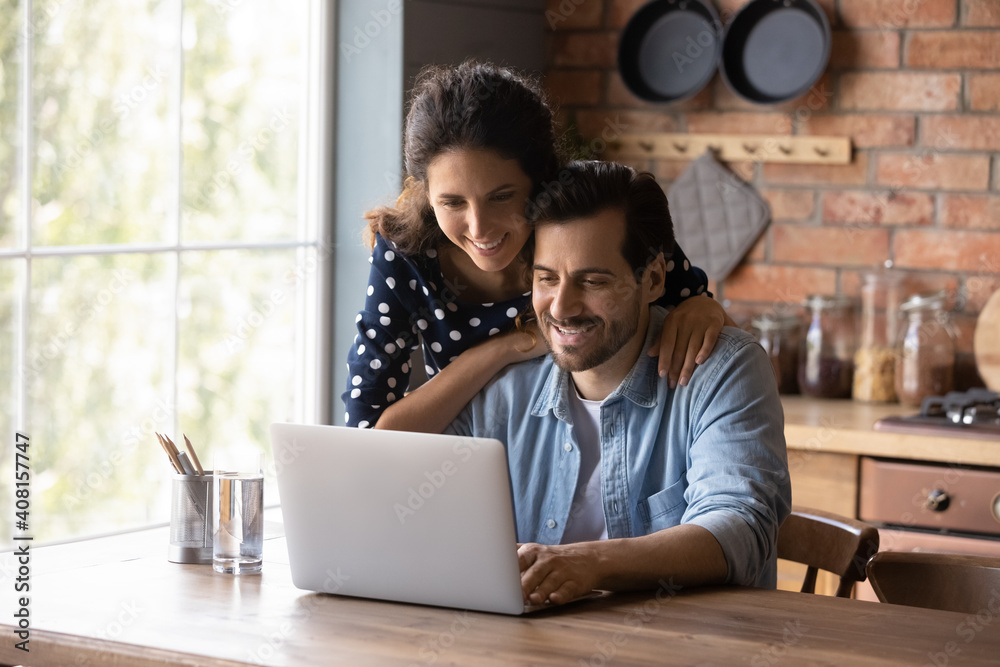 Smiling young Caucasian couple sit at table at home kitchen use laptop browsing wireless internet on gadget. Happy millennial man and woman look at computer screen shop online on device together.