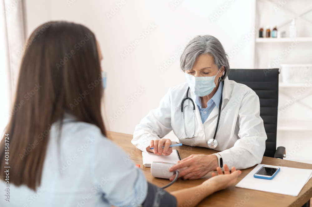 Senior mature female doctor wearing a lab coat and a face mask, using a blood pressure machine or sphygmomanometer with stethoscope to check a sick patient in the hospital during a pandemic