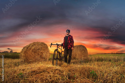 Cyclist stands with his bicycle in the field with haystacks at sunset.