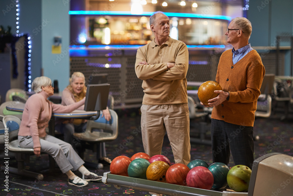 Fototapeta premium Portrait of two senior men playing bowling together with friends in background while enjoying active entertainment at bowling alley, copy space