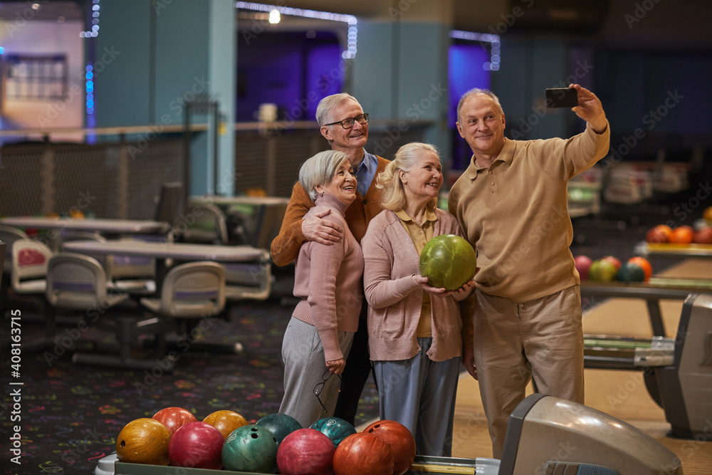 Fototapeta premium Wide angle view at group of senior people taking selfie photos playing bowling and enjoying active entertainment at bowling alley, copy space