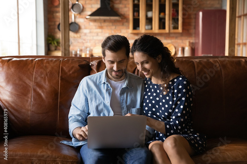 Happy young Caucasian couple rest on sofa at home look at laptop screen browse wireless internet on gadget together. Smiling millennial man and woman shopping online on computer. Technology concept.