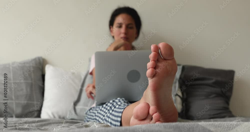 Focus on bare feet of young woman lying on cozy bed with computer on laps, working distantly from home, enjoying online shopping or communicating in social networks, feeling relaxed on weekend.
