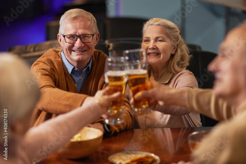 Portrait of smiling senior people drinking beer in bar and clinking glasses while enjoying night out with friends
