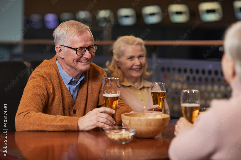 Portrait of modern senior couple drinking beer in bar and laughing while enjoying night out with friends, copy space