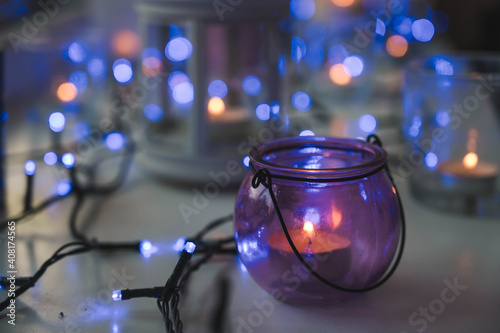 Close-up of burning tea candles in lantern and glasses on sill. Blue garland light. Hygge christmas decorations.