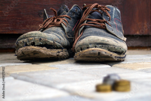 Worn boots with a lagging sole on a background of gray pavers and dark brown wood. In the foreground is a blurred stack of coins. The concept of poverty, homelessness, lack of money. Selective focus