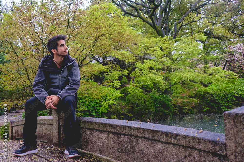 Contemplative young man in a stone bridge in a beautiful traditonal garden in a rainy day at Shitenno-ji temple in Osaka, Japan Contemplative young man in a stone bridge in a beautiful traditonal garden in a rainy day at Shitenno-ji temple in Osaka, Japan