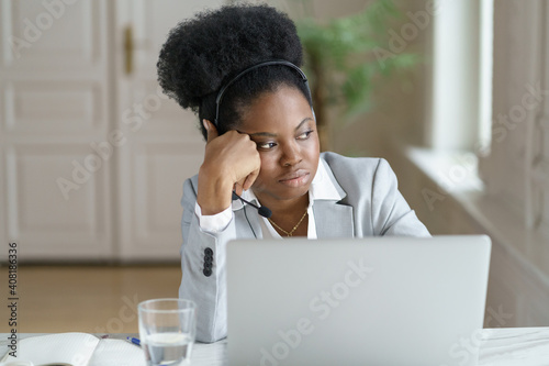 Unmotivated Afro businesswoman in blazer with headphones looking at the window, thinking or pondering, tired of monotonous work at laptop at office, dreaming about weekend vacation. Lack of motivation photo