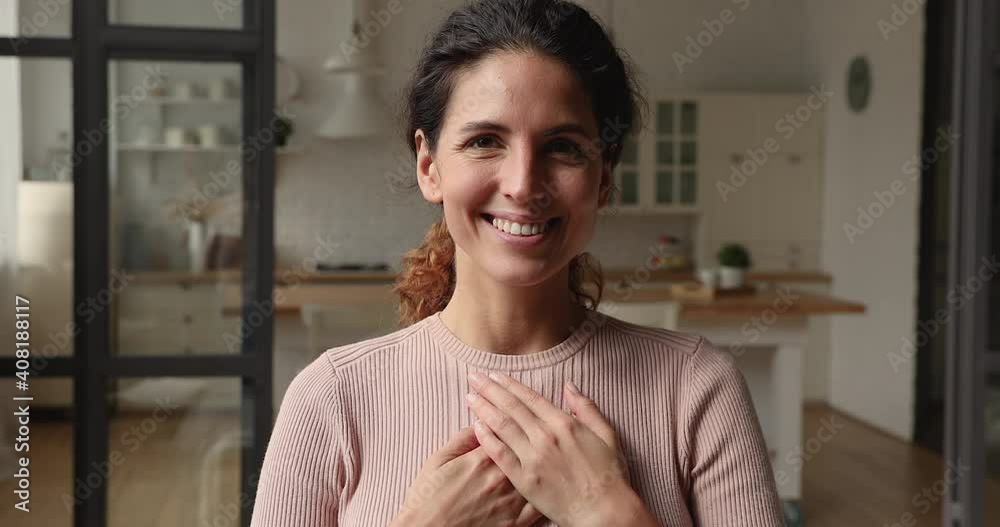 Smiling sincere kind young caucasian woman holding folded hands on heart chest, feeling deeply thankful at home. Happy inspired beautiful millennial lady showing gratitude, expressing appreciation.