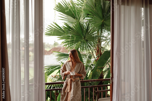 Beautiful young woman stand  on balcony terrace of jungle villa with morning coffee, wearing bathrobe. Vacation on tropical island