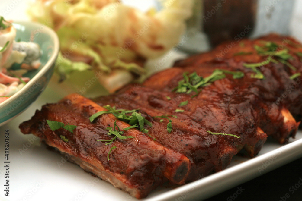 Closeup of pork ribs grilled with BBQ sauce. Tasty snack to beer on a wooden Board for filing on dark wooden background. Nearby are a baked potato and sauces for meat.