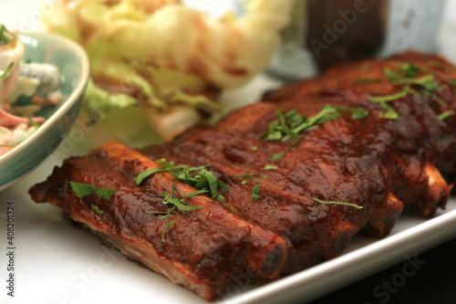 Closeup of pork ribs grilled with BBQ sauce. Tasty snack to beer on a wooden Board for filing on dark wooden background. Nearby are a baked potato and sauces for meat.