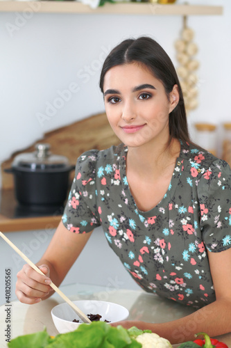 Young hispanic woman or student cooking in kitchen. Girl tasting fresh salad while sitting at the table
