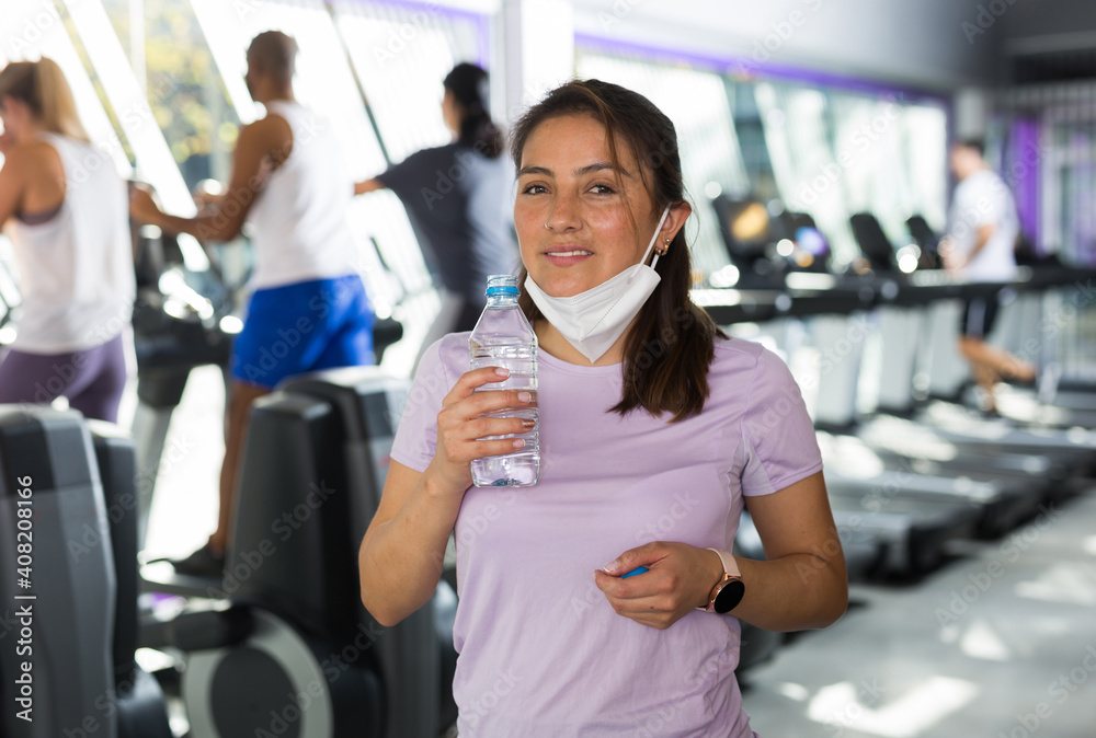 Portrait of young woman wearing protective face mask resting after workout in fitness gym holding bottle with still water