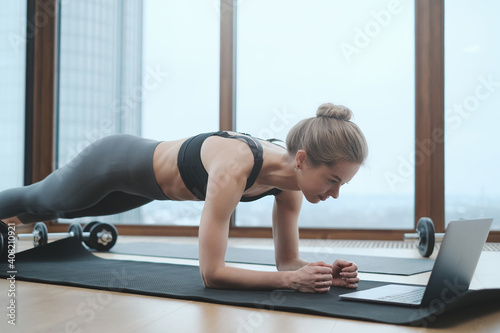 Strong caucasian woman in sportswear does physical exercise on mat looking at laptop in modern apartment.