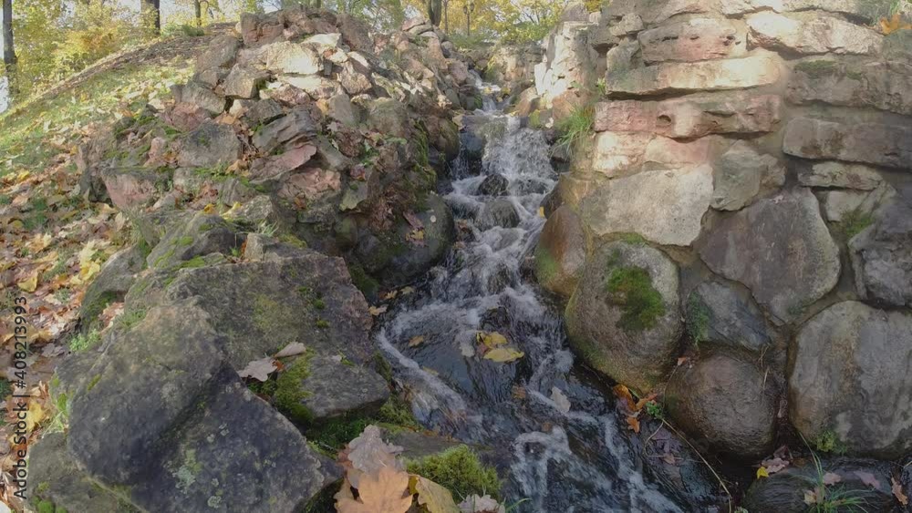 Small artificial stream runs with rapids between big brown stones lit by sun rays in autumn Riga Bastejkalns city hilly park Small artificial stream runs with rapids between big brown stones lit by sun rays in autumn Riga Bastejkalns city hilly park
