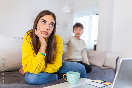 Kid anoying and disturbing exhausted and irritated mother in casual clothes holding hands on head while working at home photo