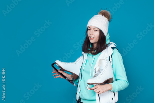 Winter hobby, ice skating. A brunette woman in a hoodie with figure skating skates on her shoulder. Studio shot on a blue background