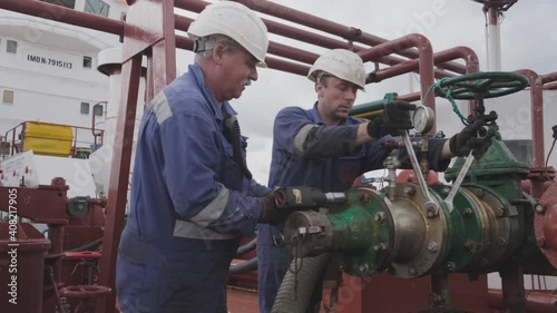 professional workers in helmets and jumpsuits repair large pipe with silver spanners under red pipelines against cloudy sky