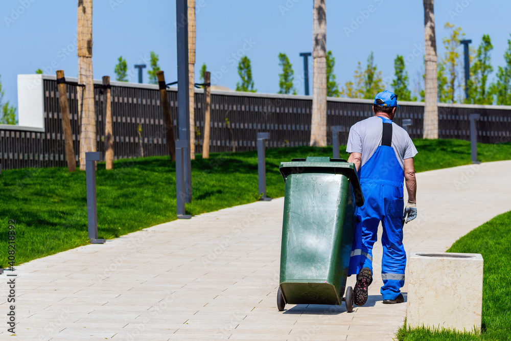 Janitor rolls trash can on wheels. Cleaning service in modern park