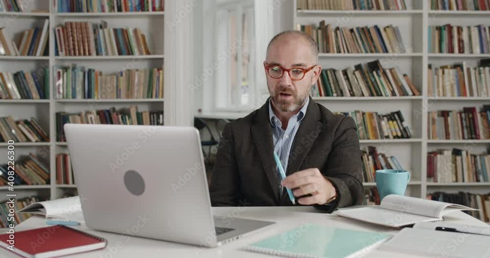 Crop view of handsome man in glasses talking and holding pen while looking at laptop screen. Male teacher in suit having online lesson while sitting in room full of books Crop view of handsome man in glasses talking and holding pen while looking at laptop screen. Male teacher in suit having online lesson while sitting in room full of books
