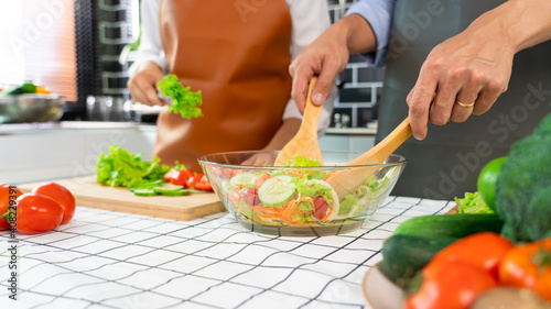 Happy couple having fun standing in kitchen at home preparing vegetable salad husband and wife vegetarians chop vegetables prepare for dinner in loft kitchen at home.