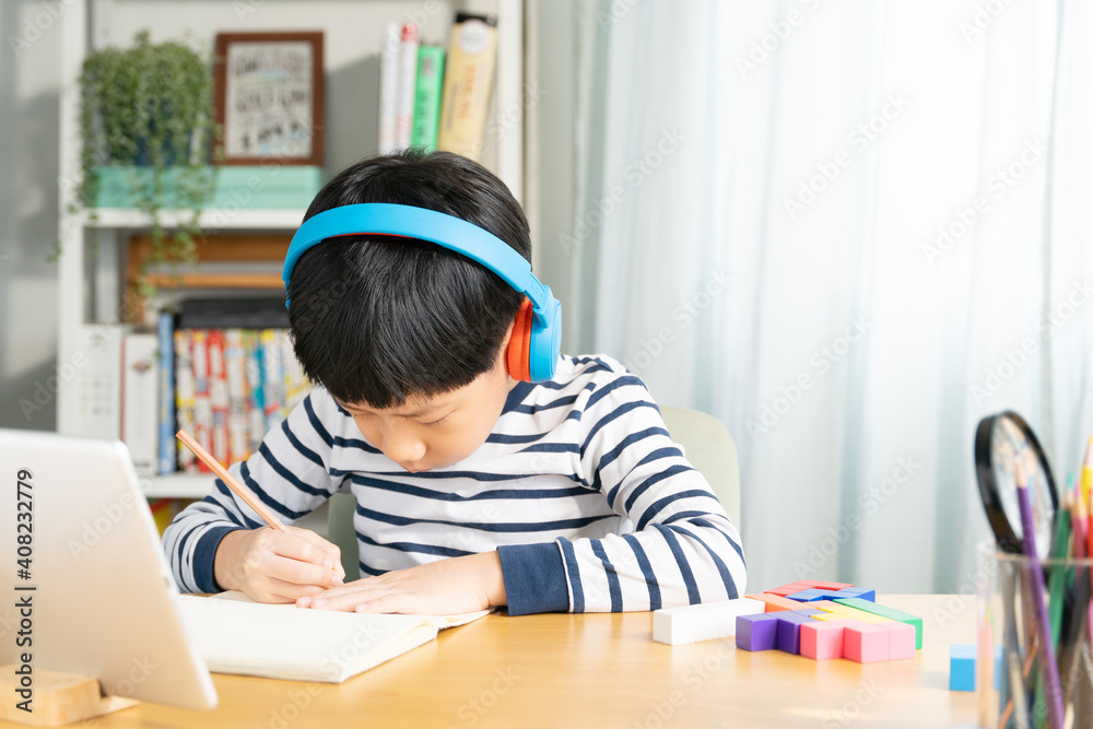 Portrait of smart Asian student boy with headphone, write, study at home with tablet computer in online classroom during Covid-19 pandemic lockdown. New normal in technology, education, homeschool