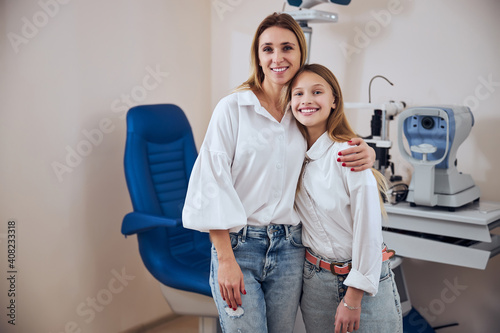 Happy smiling woman and teenager girl standing in front of the different modern machine in optician center