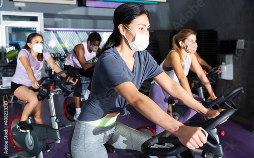Portrait of young adult woman training on cycling machine in gym, all people wearing face masks for disease protection