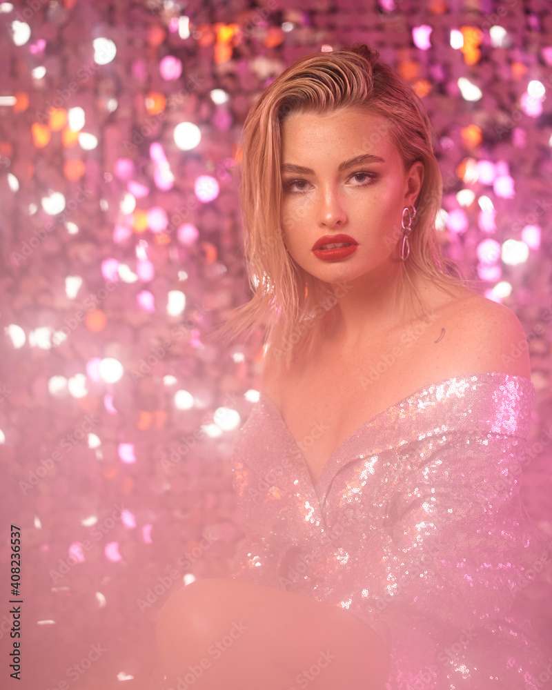 Fashionable partly defocused blurred studio portrait of young sexy blonde woman with makeup and middle length straight hair in shining silver dress against glamor sequins background. Pink light