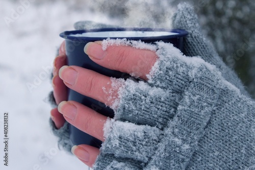 Woman holding a mug in her hands. The gloves are covered in snow. It`s freezing.Side view of frozen hands of a young woman holding a mug.