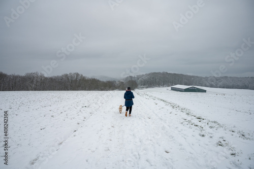 eine Frau läuft mit einem kleinen Mischlingshund durch eine verschneite Landschaft im Winter 