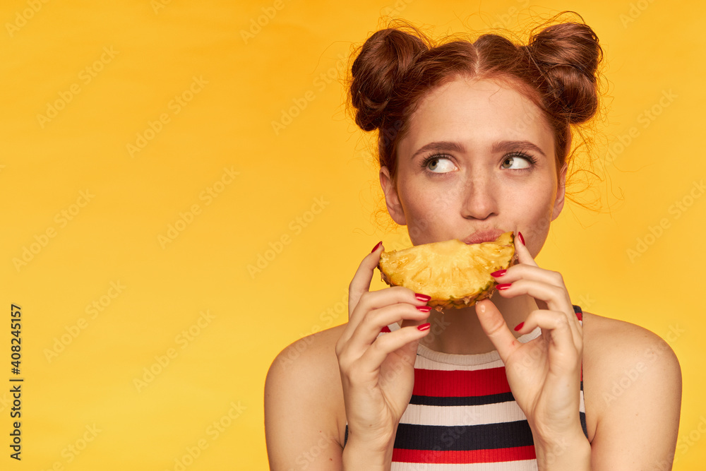 Young lady, pretty ginger woman with two buns and healthy skin. Wearing striped tank top and biting slice of a pineapple. Watching to the left at copy space, isolated closeup over yellow background Young lady, pretty ginger woman with two buns and healthy skin. Wearing striped tank top and biting slice of a pineapple. Watching to the left at copy space, isolated closeup over yellow background