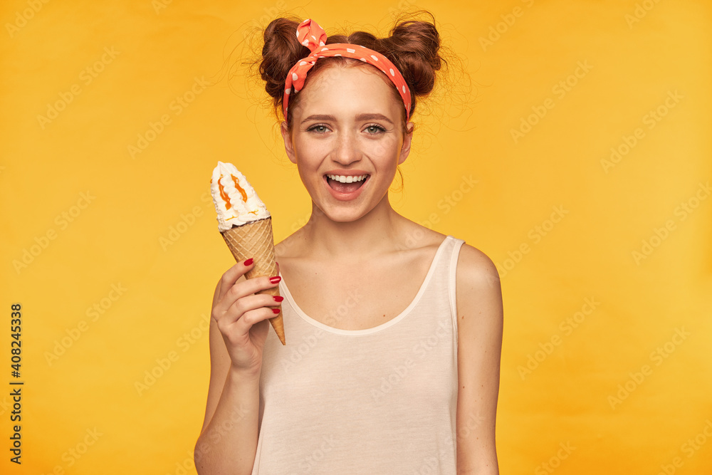 Teenage girl, happy looking red hair woman with two buns. Wearing white tank top and red doted hairband. Holding ice cream and have a big smile. Watching at the camera isolated over yellow background Teenage girl, happy looking red hair woman with two buns. Wearing white tank top and red doted hairband. Holding ice cream and have a big smile. Watching at the camera isolated over yellow background